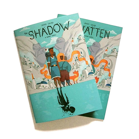 A photo of the printed English and German editions of my comic „The Shadow“, showing the two main characters in front of a city in a valley. They're standing (on the shore?) of a lake. There's a shadowy creature instead of the reflection of one of the main characters in the water.