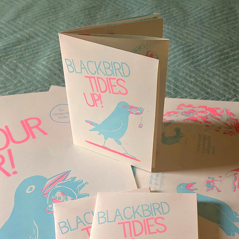 Photo of a riso-printed mini folding zine in bright pink and blue called „Blackbird Tidies Up!“, showing a giant blackbird with a car in it's beak. There's the inside of the poster next to it, showing blackbird with the Monopoly guy in it's beak.