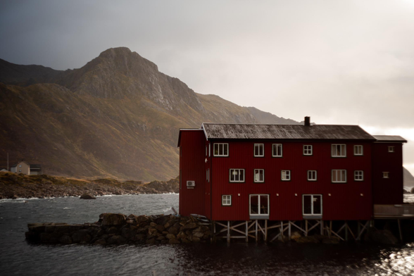 An old wooden red painted building built on poles on the shore of the sea, mountain in the background