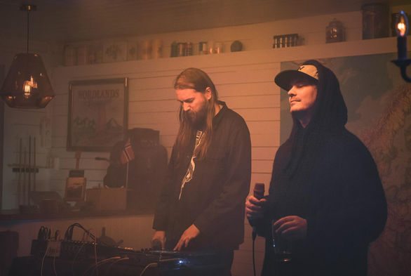 Kyber with long hair and hoodie, and Martin Massiv wearing a cap and hoodie, inside in a cozy pub with a bunch of electronics