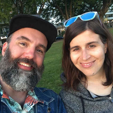 A selfie of me and my wife Candice in a park. I'm wearing a baseball hat, full beard, and a Hawaiian shirt with a jean jacket. Candice has sunglasses on her head and is wearing a grey hoodie.