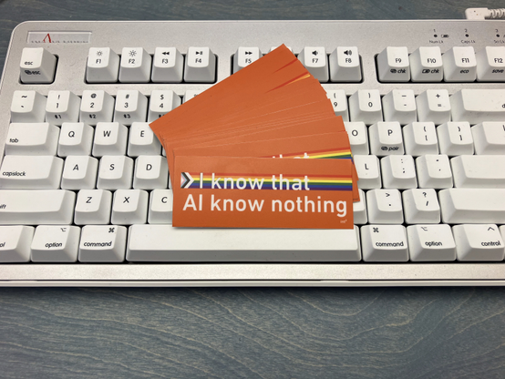 An orange sticker with the text "I know that AI know nothing" embossed. The progressive pride flag is used as the background for the first line.

The sticker rests on a white Realforce R3 keyboard (mac Edition)