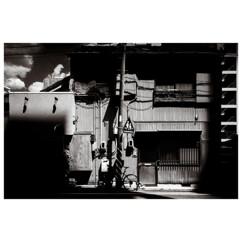 A black-and-white photo of a quiet Nagoya street scene. The building façade is made of corrugated metal, lit by sharp sunlight that casts deep shadows from nearby structures. A bicycle leans against a utility pole in front, next to a pedestrian crossing sign. The textures of metal, shadow, and light create an unexpectedly warm, nostalgic feeling despite the industrial materials.