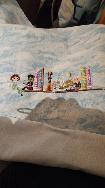 Light blue evenweave fabric, with cross stitch motifs. from left to right: Mary Shelley, Frankensteins monster, a purple then blue book, the tower from lord of the rings, a waterfall, white mountain, hobbithut, J.R.R Tolkien a gren and a purple book