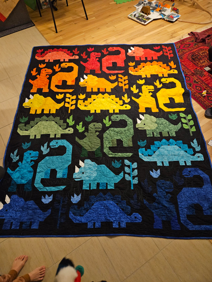 Modern patchwork quilt. Black baggrund with 6 of each dinosaur,  triceratops,  stegosaurus,  T-rex and Brachiosaurus, some vines and 2 other types of fauna.
Arranged from top to bottom in vibrant red, orange,  yellow, green, light blue and dark blue