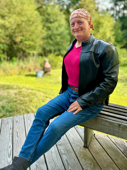 The thumbnail of this photo likely showed either my face or my boobs, depending on your app. I am sitting on a grey wooden bench on my porch and looking almost directly at you with a friendly and relaxed smile. Behind me is green grass, shrubs, and trees. 
Starting at the top, on my head I am wearing a headscarf the color of a capuchin monk’s robes with a simple white floral print. Sixty-plus years of smiling have given me a fan of wrinkles between my eyes and temples. I have just a hint of dimples in my cheeks, and a strong chin cleft that puts Kirk Douglas’s little chin dimple to shame. I am wearing a Euro-style black leather motorcycle jacket (with the shoulder and elbow armour still in place, oops!) over a deep pink-red top. The fabric-covered mounds of my training breasts are peeking out from the unzipped jacket, showing you what my body hopes to become. Below the the waist I am wearing stretch jeans that are a size too big to stretch and a brown leather belt.  My legs are crossed slightly at the ankles, and you can just see the tops of apair of charcoal grey suede boots. My legs and waist are slender, with pleasant padding on my hips (another bit of sympathetic magic). My left hand rests on my thigh, fingers slightly spread. The pinky nail is painted pink, and a worn golden wedding band adorns the ring finger. The index finger is suspiciously close to my crotch, which shows no sign of a bulge. Just folds of denim from the oversized jeans.