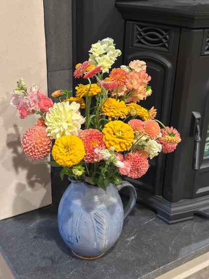 Flowers grown by a local friend and arranged in a big blue pitcher by my lovely wife. Loads of dahlias and some freesia and other blooms form a riot of pink and orange and yellow. The pitcher is resting on a granite hearth next to a cast iron pellet stove.