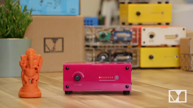 A bright pink plastic box with a knob and a row of lights on the front is sat on a table.  Next to it is an orange 3D print of Ganesha.

The lights on the box illuminate in turn and when the last one lights up the box announces "Ready!".

A hand places the Ganesha print onto the box, and the box then begins playing sitar music
