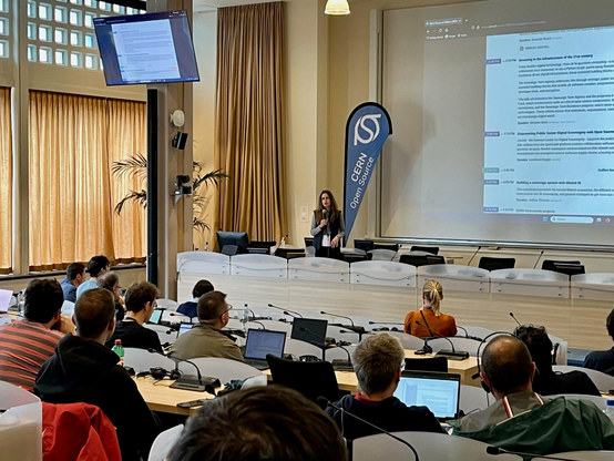 Adriana Groh speaking in front of a large audience with a "CERN Open Source" banner next to her.