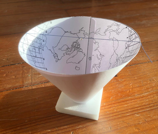 A paper cone printed on the inside, placed on a wooden floor, and under sunlight. We can see a grid and a map printed. There's a thread crossing the upper opening, holding a very small bead right on the axis of the cone.