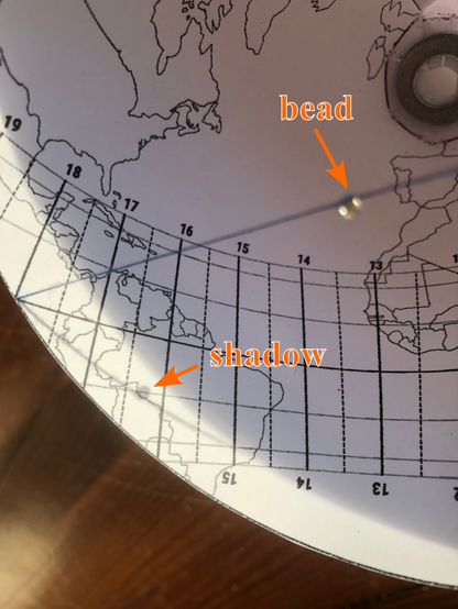 Close view from above: the bead, its shadow on the grid lines and the map. The shadow says its approx. 16h20 (4:20 pm), and its near the Brazil-Bolivia border on the map: at the time of this photo the Suns was directly overhead at this place.