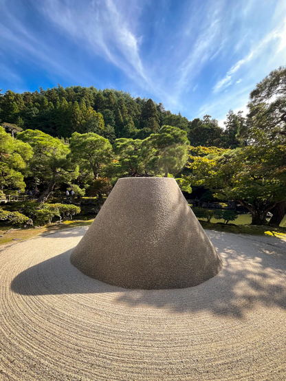 Un jardín zen (gravilla rastrillada) formando círculos alrededor de un cono de piedra sin pinta que recuerda vagamente el monte Fuji. Detrás, los árboles del jardín.