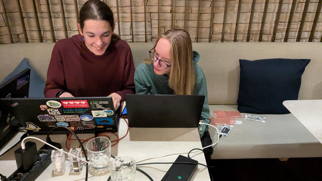 two people sitting at a table with two laptops, cables, and phones hacking together.