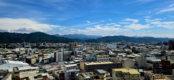 Das Bild zeigt eine Stadtansicht, aufgenommen an einem klaren Tag mit weitem Blick über eine urbane Landschaft.

Im Vordergrund sieht man zahlreiche Gebäude unterschiedlicher Höhe – überwiegend flache Wohnhäuser, einige mittelhohe Büro- oder Apartmentgebäude. Die Dächer wirken teils modern, teils älter, viele mit typischen Blech- oder Betonkonstruktionen.

Im Hintergrund erstreckt sich eine beeindruckende Bergkette mit mehreren dicht bewaldeten Hügeln, die in verschiedenen Blautönen in der Ferne verschwimmen – ein Effekt, der auf die Dunst- oder Nebelschichten in der Sommerluft hindeutet.

Der Himmel ist weitgehend klar, leicht bewölkt, mit zarten, weißen Wolkenstreifen und einem hellen Blau – ein typischer Anblick tropischer oder subtropischer Städte an einem sonnigen Tag.

Insgesamt vermittelt das Bild eine ruhige, helle und weite Atmosphäre mit einem harmonischen Übergang zwischen Stadt und Natur.