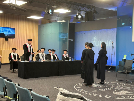 Part of the ceremony. The committee in robes are sitting behind a table, while one member stands up and speeches. Freshly minted doctor and her paranimphs are standing in front of them, with their backs to the viewer, listening. All are dressed in black. TU Delft logo in background, coat of arms on floor.