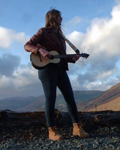 1) A photo of me playing a baritone ukulele outside in the Scottish hills. My uke has a spruce top and rosewood neck, back and sides, and is supported with a white and yellow strap. I am wearing a leather jacket, jeans and boots. The hills are laid out going up a glen behind me, and are a mixture of grass and forestry. It is a sunny day with white clouds.