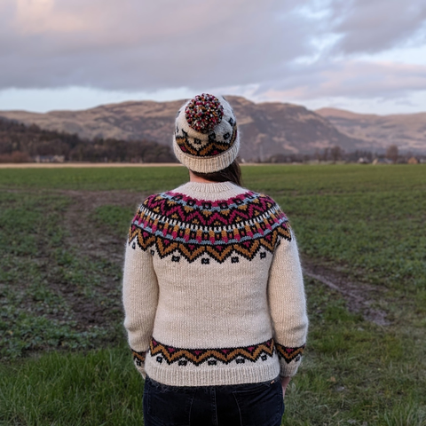 2) A photo of me facing away from the camera, wearing a handknit jumper and matching hat. The jumper is a lopi yoked jumper, with main colour white and a colourwork yoke, hem and cuffs with bold geometric shapes in yellow, pink and blue, all outlined in black. The hat has the same pattern around it, and a pompom on top made of all the yarns. It is a late-winter Scottish day and in the background are some low hills.