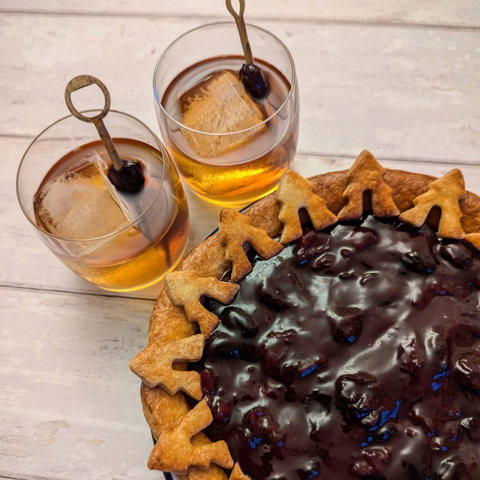 3) A close-up square photo of a pie, with two cocktails to its top left, on a white wooden background. The pie has a golden brown crust decorated with little pastry fir trees all around, surrounding a dark-red cherry topping. The cocktails are two whisky-coloured drinks in rocks glasses, each with one large ice-cube and a cherry garnish on a golden cocktail stick.