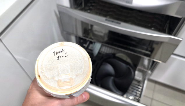 Das Foto zeigt eine Nahaufnahme eines Getränkebechers mit Deckel, auf dem handschriftlich „Thank you :)“ steht. Der Becher wird über einer geöffneten Schublade eines Geschirrspülers oder Geschirrtrockners gehalten, in dem sich schwarzes Geschirr und Küchenutensilien befinden. Die Szene spielt in einer modernen Küche mit hellen Arbeitsflächen und Edelstahlgeräten.