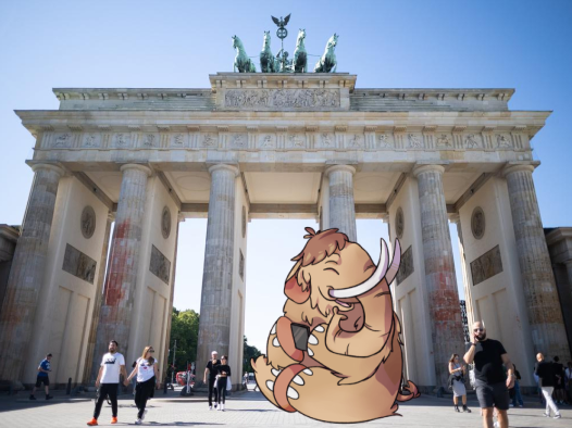 Das Bild zeigt das Brandenburger Tor in Berlin im Sommer vom Osten aus fotografiert. Vor dem Tor sind einige Menschen zu erkennen, und zudem wurde das lachende Mastodon-Maskottchen in das Bild integriert. 