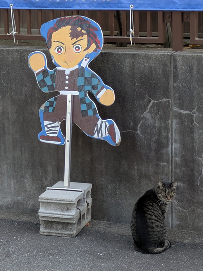 「鬼滅の刃」の竈門炭治郎を模した飛び出し坊や看板と横に座る野良猫。町中のスナップ。