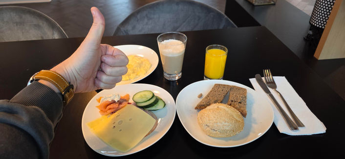 Auf dem Foto sieht man ein reichhaltiges Frühstück auf einem schwarzen Tisch – und eine Hand, die mit einem Daumen nach oben zeigt, was eindeutig Zustimmung signalisiert.

Zu sehen sind:

Ein Teller mit Rührei

Ein Glas Milchkaffee und ein Glas Orangensaft

Ein weiterer Teller mit Käse, Wurst, Lachs, Gurkenscheiben

Ein Teller mit Brötchen und einer Scheibe Vollkornbrot

Besteck auf einer Serviette rechts daneben

Es wirkt wie ein klassisches Hotelfrühstück – ordentlich angerichtet, frisch und offenbar absolut „Daumen-hoch“-würdig.

