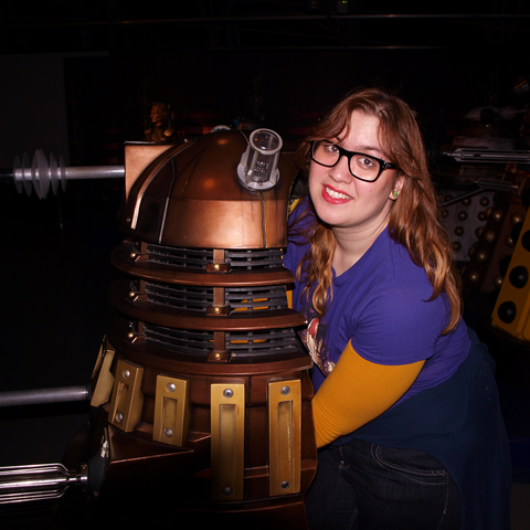 Rosie is a plus size Dutch Indonesian woman with a light to medium skin tone and brown eyes. In this photo she has ginger hair. She is wearing black frame glasses, a dark pink lipgloss and green earrings. She is wearing a purple Doctor Who t-shirt with Donna Noble on it, and underneath there's a yellow long sleeved shirt. Rosie is controlling a bronze coloured Dalek at the Doctor Who Experience in Cardiff, Wales. In the background you can spot a whole collection of Daleks. Rosie is looking at the camera and is smiling.