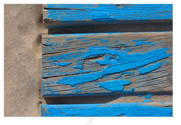 A close-up photo in landscape format. The view is directly from above onto a wooden board of a jetty lying on the sand of a beach. The boards are painted in a fresh blue color. Above and below are two more wooden planks, but they are only partially visible. The weather has already caused the paint to peel off in places. The light-colored sand can be seen between the boards.

AI disclaimer: Using my work, its meta data, written or derived description to create media with or train AI based systems is prohibited.