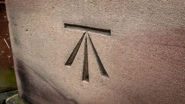 A non-print example.

The most common form of the symbol, although other variants exist. It's carved into a smooth block of stone on the side of a building or monument plinth, and it looks like a capital T with an upside down V overlaid onto it so that the two angled lines from the V come together with the T's vertical stroke to meet its horizontal stroke at a single point. All the bottom lines taper toward that point.  Looking at it another way, it's a horizontal line with an arrow pointing at it, saying "here, this level!"

They were used for marking height reference points during various surveys of the British isles.

Photo by Mike Taylor on geograph.org.uk, CC:by