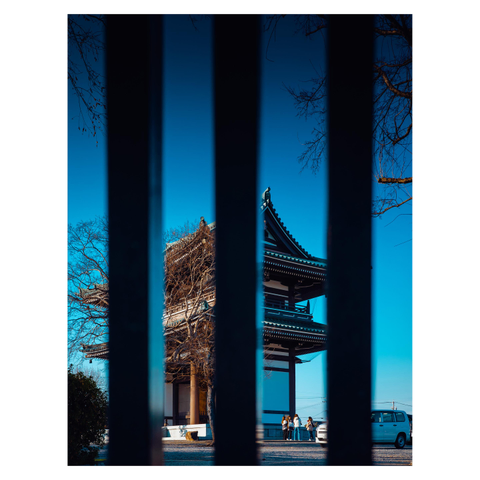 A color photo of a temple at Kakuōzan in Nagoya, viewed through vertical black bars in the foreground. The temple’s wooden architecture and sweeping tiled roof stand out against a vivid blue sky. A few visitors and a small white van are visible near the entrance.