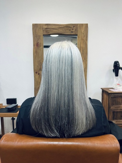 背中を向けて椅子に座る人物の後ろ姿。肩下まであるストレートのロングヘアは美しいシルバーグレーで、均一に艶がある。正面には木枠の鏡があり、美容室の落ち着いたインテリアが背景に写っている。