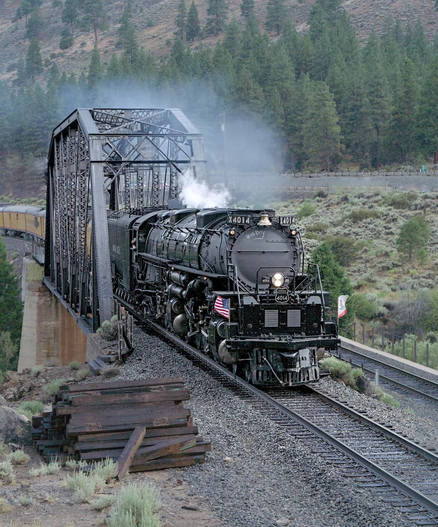 A color portrait image of a very large steam engine that has just exited a metal bridge structure over a river. Forest is seen in the background. The engine is approaching the viewer, exiting the frame at the lower right. A big brass bell is seen at the top of the tubular black boiler. A small US flag is on the right corner of the engine and a small California flag is on the left. A small pile of railroad ties lies on the gravel railroad bed in the lower left.