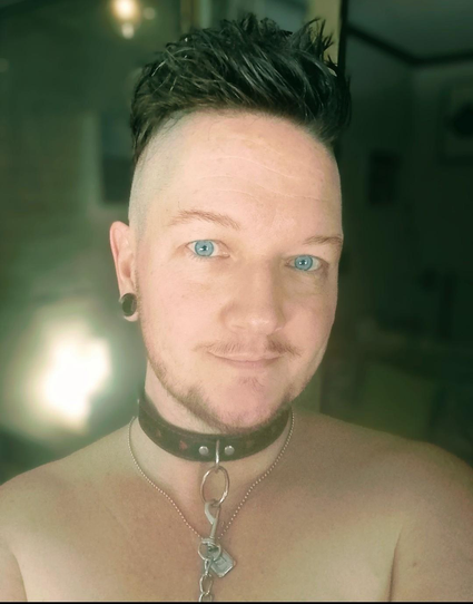 White trans man with dark facial hair and piercing blue eyes. He looks into the camera, with a slight smile. He wears nothing but a day collar(silver necklace with "B" pendant), and another collar that's back with red hearts on it. The collar is tight around his neck, and has a leash. The pic is taken from the chest up.
