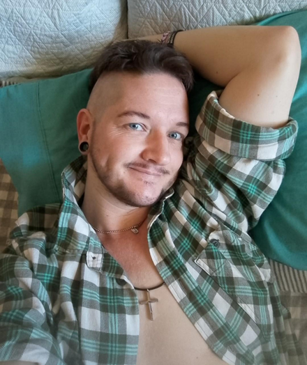 White trans man with dark hair and facial hair and piercing blue eyes. He smiles as he looks into the camera. He wears an open green, grey and white plaid shirt, with sleeves rolled up, and has one arm behind his head as he reclines on a blue cushion. He has a slightly suggestive smile that shows his dimples. He wears a cross around his neck, and another pendant.