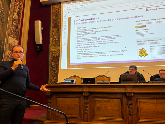 Photo prise dans la salle du Sénat de l’université Paris Cité, reconnaissable à ses tentures. Je tiens le micro à gauche et sur la droite se trouvent à La Tribune le directeur de l’UNIF, Dominique Bascle et Gilles Roussel président de son assemblée général. L’écran au dessus d’eux affiche une page de présentation où l’on reconnaît en bas à droite le logo de bonfire.