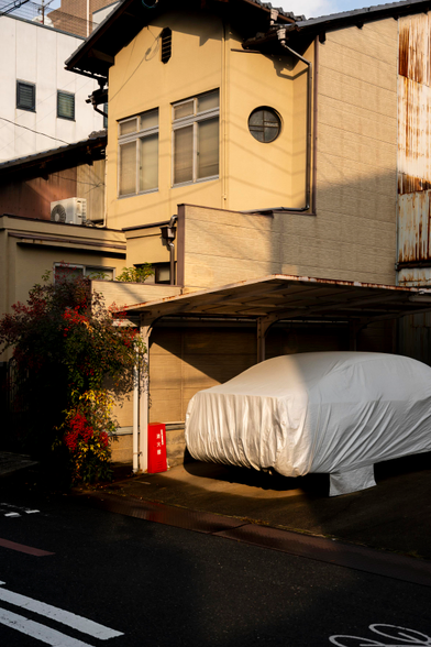 A car wrapped in plastic is parked under a semi-covered parking spot. There’s a metal red box next to it in the edge of the wall, which has some colorful leafy bushes at its end, next to the street. Behind the spot there’s a two level house, in light yellow. And a few more houses behind. They’re all very narrow. The scene is basked in a nice golden sunlight.