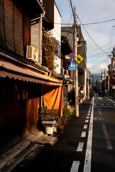 A narrow street is seen, with many houses along its edge. The nearest house has an orange awning shielding its wooden plank walls from the elements. Along the street there’s an electric pole with some road signs attached, including two caution with pedestrian signs and a no parking sign. Further along the street there’s a cyclist, and a car further down still. Power lines crisscross the street along its distance, and the sky is a mix of light blue and grey. The street is cast in a golden light.