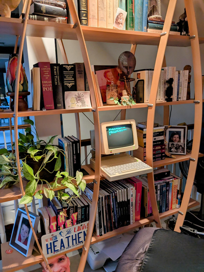 Picture of my shelf in my office with books, a plant, knickknackery, and a vintage Apple IIc that still works after 40 years.