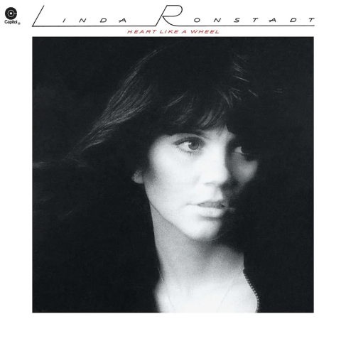 A black and white photograph of a woman on an album cover. The background is completely black, accentuating her facial features. Her head is turned slightly to the right, with her gaze directed off-camera. She has voluminous, wavy hair that partially covers her forehead. Her expression is serene and introspective. The upper portion of the image contains text in a stylized font. The cover is designed with a stark contrast between the lightness of her face and the surrounding dark background.

Transcribed Text:

Capitol LINDA RONSTADT HEART LIKE A WHEEL