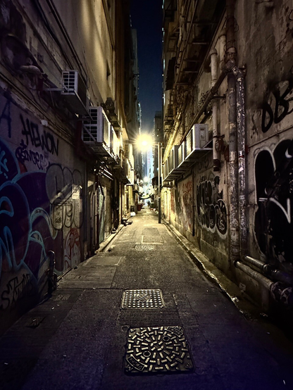 夜の細い裏路地。両側の古びた建物の壁には落書きが広がり、外付けエアコンや配管がむき出しになっている。頭上から黄色い街灯が路地を照らし、奥へと続く細長い通路の先にかすかな灯りが見える。舗道には複数のマンホール蓋があり、静まり返った都会の裏側の雰囲気が漂っている。