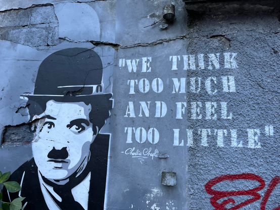 Graffiti, a stencilled black and white depiction of Charlie Chaplin with moustache, bowler hat. Next to him is stencilled the phrase “WE THINK TOO MUCH AND FEEL TOO LITTLE”.