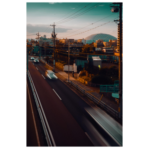 A color photograph taken from an elevated position overlooking a road at sunset. Cars move quickly, creating soft motion blur along the lanes. Power lines and utility poles crisscross the scene. In the distance, the large domed roof of Nagoya Dome rises above low buildings, lit warmly by the setting sun under a pale blue-green sky.