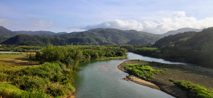 Eine Landschaftsaufnahme zeigt ein dicht bewachsenes, tropisches Tal bei Sonnenuntergang. Die tief stehende Sonne rechts taucht den Himmel in warme Gold- und Orangetöne und sendet helle Strahlen über die Szene. Die Baumkronen in der Schlucht sind in tiefes Grün getaucht, während am oberen Horizont mehrere Häuser mit hellen Dächern sichtbar sind.