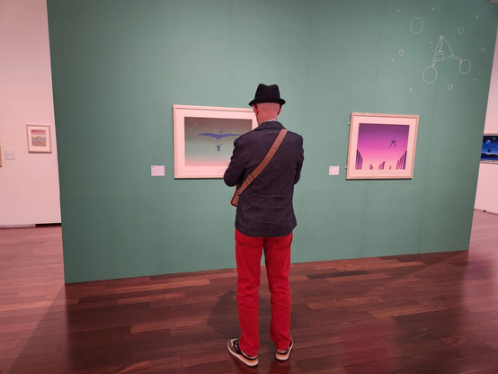 A man wearing a dark blue denim hat, dark blue denim blazer, red denim pants, and sneakers is standing in an art gallery, looking at framed artworks by Folon displayed on a green wall. The paintings feature surreal scenes with minimalist, colorful backgrounds. The gallery has wooden flooring and clean, modern decor.