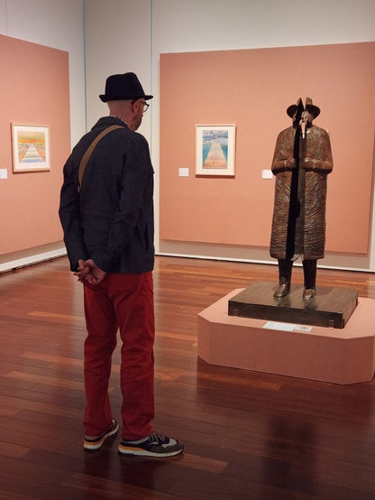 A man wearing a dark blue denim hat, dark blue denim jacket, red denim pants, and sneakers is observing a bronze sculpture by Folon of a person wearing a trench coat and hat. The sculpture is split vertically down the middle. Surrounding the sculpture are framed artworks mounted on a peach-colored wall.