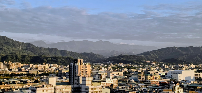 Eine Weitwinkelaufnahme einer weitläufigen Stadtlandschaft unter einem hellblauen Himmel mit lockeren Wolkenformationen. Im Hintergrund erstreckt sich eine markante, bewaldete Bergkette, die klar gegen den Horizont abgegrenzt ist. Die städtische Bebauung im Vordergrund besteht aus einer Vielzahl von Gebäuden mit flachen Dächern, auf denen häufig Wassertanks und technische Installationen zu sehen sind. Das Sonnenlicht beleuchtet die Fassaden der Gebäude im mittleren Bildbereich und hebt deren ockerfarbene und weiße Töne hervor.