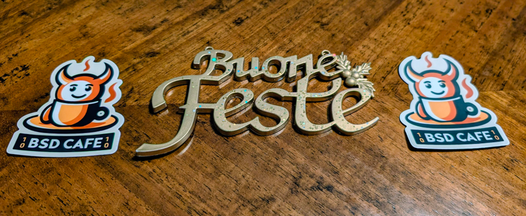 Wooden tabletop with a glittery gold "Buone Feste" (Happy Holidays) decoration in the center, flanked on both sides by identical BSD Cafe stickers.