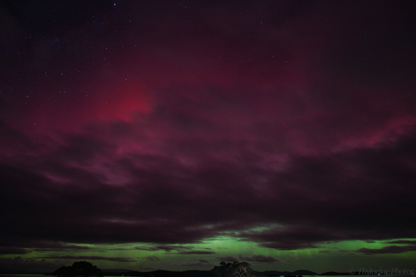 An aurora fills most of the frame, with stark grey clouds filtering the red/pink glow above the typical green, closer to the horizon.  Distant water reflects the green sky, and tree lined hills are just visible in the distance.