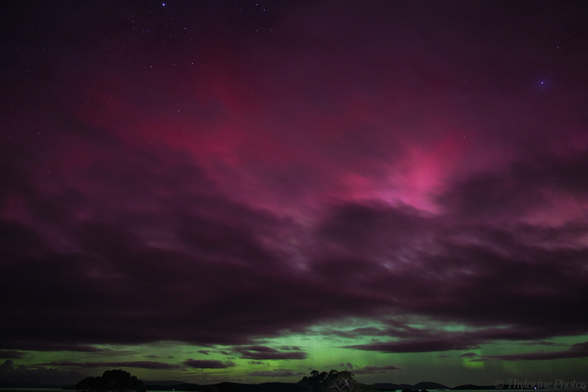An aurora fills most of the frame, with stark grey clouds filtering the red/pink glow above the typical green, closer to the horizon.  Distant water reflects the green sky, and tree lined hills are just visible in the distance.