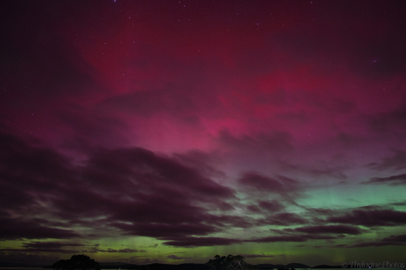 An aurora fills most of the frame, with stark grey clouds filtering the red/pink glow above the typical green, closer to the horizon.  Distant water reflects the green sky, and tree lined hills are just visible in the distance.  The cloud is starting to thin out compared to the previous images, with most of the top half of the frame a reddish pink glow.